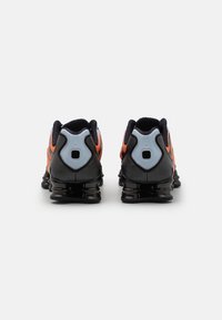 Svarta och grå sportskor med orange detaljer, som har en strömlinjeformad design, texturerat material och en distinkt form på baksidan.