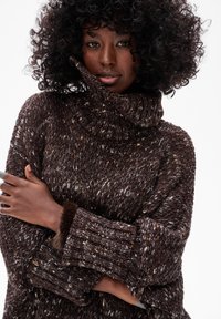 Kobieta o kręconych włosach, ubrana w gruby brązowy sweter z wrabianym wzorem, skrzyżowała ramiona i patrzy bezpośrednio przed siebie na jednolitym tle.