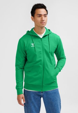 Mężczyzna w zielonej bluzie z kapturem zapinanej na zamek błyskawiczny z logo na piersi i niebieskich jeansach, stoi z jedną ręką w kieszeni na jednolitym tle.