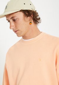 Sweatshirt de couleur pêche avec un col rond côtelé et un détail de logo doré. Porté avec une casquette beige clair et une boucle d'oreille colorée.