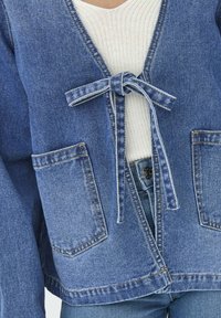 Jeans jakna, svijetlo plava, s vezanjem sprijeda, dvije prednje džepove i opušteni kroj. Nosi se preko bijelog rebrastog topa.