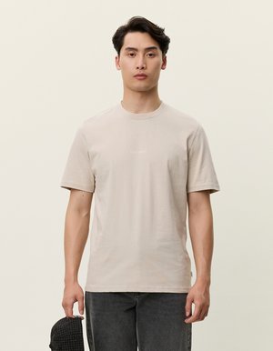 Beige T-Shirt mit kurzen Ärmeln aus weichem Stoff, mit einem einfachen Rundhalsausschnitt und einem minimalistischen Frontlogo. Getragen mit dunklen Jeans.