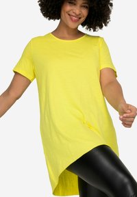 Tunic gialla a maniche corte con scollatura rotonda e design orlo alto-basso. Realizzata in tessuto leggero, abbinata a leggings neri.
