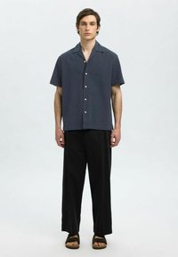 Kortsleeve, teksturert marineblå skjorte med hvite knapper, kombinert med høy midje svarte bukser og mørke sandaler, på en nøytral bakgrunn.