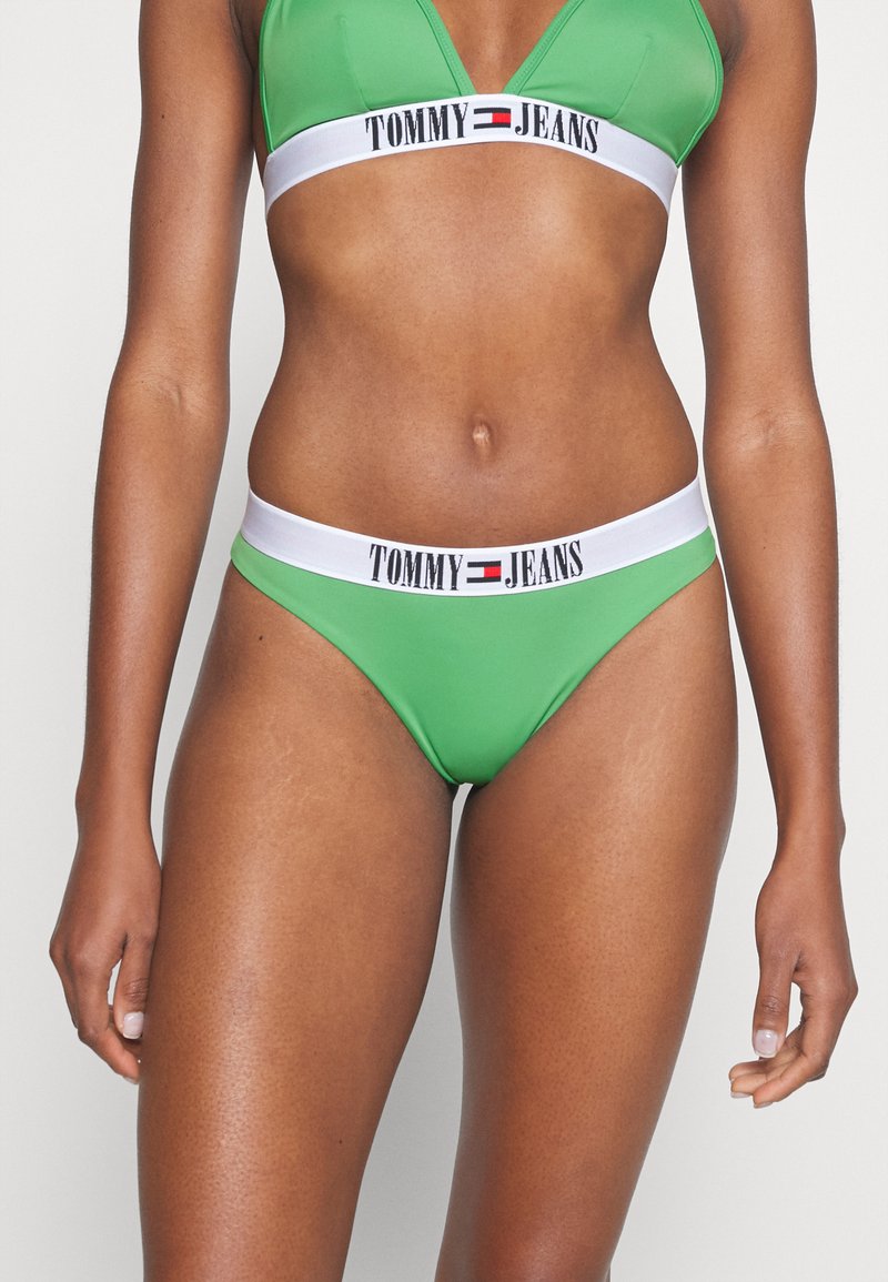 Zelená bikiny set obsahující trojúhelníkový top a odpovídající nízko vystřižené kalhotky. Elastický pás má bílý pruh s nápisem "TOMMY JEANS" vytištěným černou barvou.