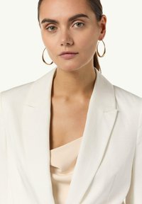 Weißer Blazer mit Reverskragen, kombiniert mit einem beige Satinoberteil. Runde goldene Creolen ergänzen das Outfit. Glatte Texturen.