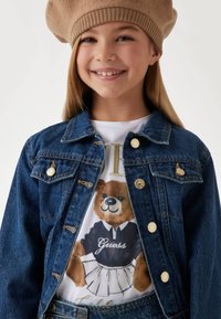 Jeansjacka med guldknappar, med en vit t-shirt som visar en björn i en marinblå skjorta och vit kjol med texten "Guess".