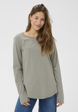 Mlada ženska z dolgimi rjavimi lasmi, ki nosi zlata obročasta ušesa, svetlo sivo majico z dolgimi rokavi in modre kavbojke, nežno se smehlja.