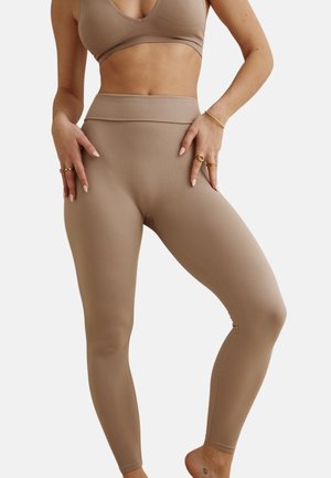 Femme portant des leggings côtelés beige taille haute et un soutien-gorge de sport assorti, debout les mains posées sur les cuisses, pieds nus sur fond blanc.