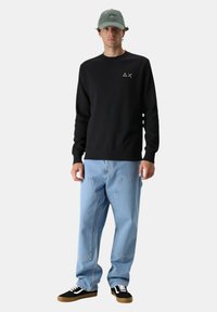 Giovane uomo che indossa una felpa nera con un piccolo logo, jeans baggy azzurri chiari, sneakers nere e un cappellino da baseball grigio, in piedi su uno sfondo bianco.