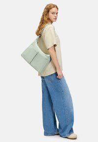 Bolso de hombro verde claro en material suave, con un bolsillo delantero con cremallera y una correa ancha, llevado por una persona con una camisa beige y pantalones anchos.