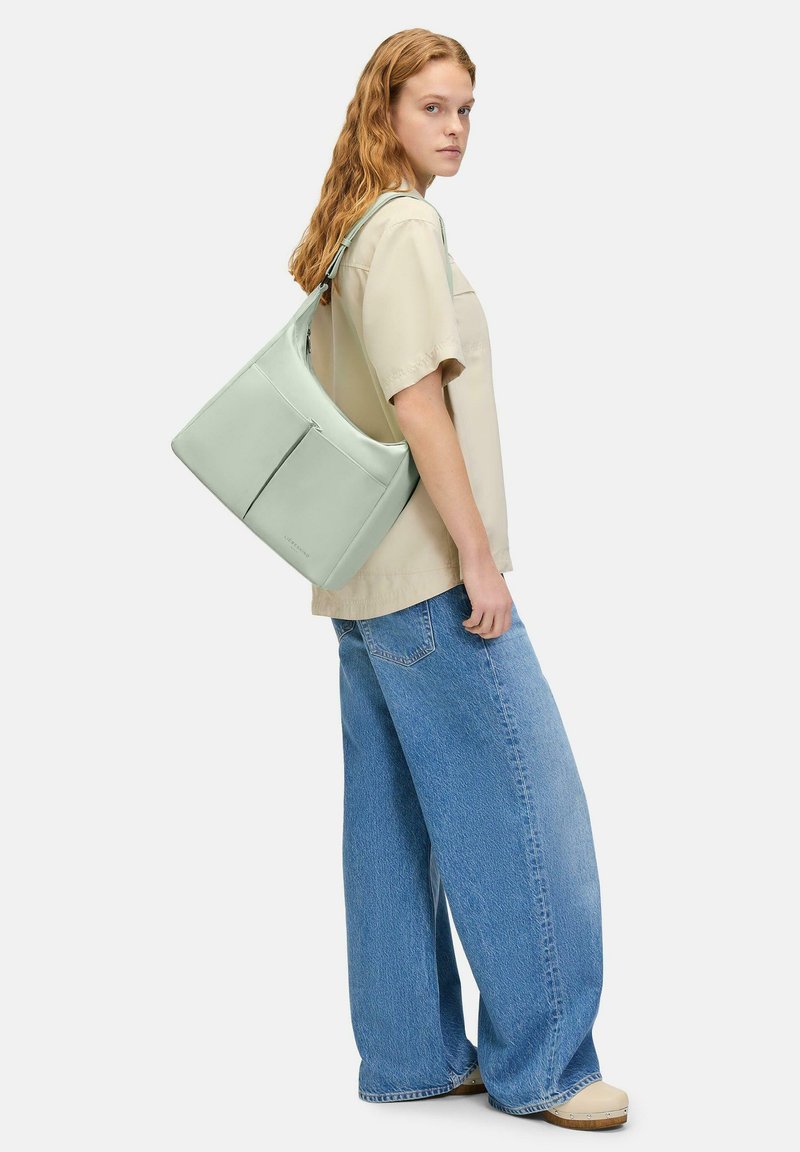 Bolso de hombro verde claro en material suave, con un bolsillo delantero con cremallera y una correa ancha, llevado por una persona con una camisa beige y pantalones anchos.