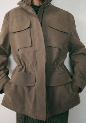 Bruine casual jas met vier klepzakken aan de voorkant en een elastische taille, gedragen over een groen geribd truitje.