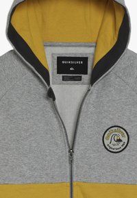 Šedá a žlutá mikina na zip s černým lemováním uvnitř kapuce, nášivka loga Quiksilver na hrudi a štítek značky uvnitř límce.