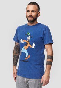 Sininen puuvillainen t-paita, jossa on sarjakuvahahmo Goofy leikkisässä asennossa. Lyhyet hihat, pyöreä pääntie ja rento mitoitus.