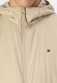 Beżowa kurtka z kapturem, częściowo zapinana na zamek z przodu, z małym prostokątnym logo w kolorach czerwonym, białym i niebieskim na piersi.