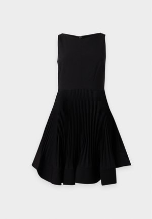 Femme portant une robe noire plissée sans manches et des ballerines noires à bride arrière, tenant un grand sac à main en cuir noir sous le bras.