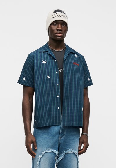 Άνδρας που φοράει λευκό σκουφί με μαύρο σχέδιο, μπλε πουκάμισο με κοντά μανίκια σε καρό σχέδιο με λευκά μπαλώματα κουνελιού, μαύρο μπλουζάκι με τύπωμα και σκισμένο μπλε τζιν.