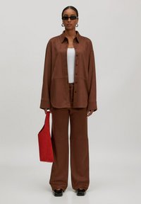Vrouw in een oversized bruine blouse en broek, witte tanktop, zwarte schoenen, zonnebril, oorbellen in ringen, met een rode tas aan haar zijde.