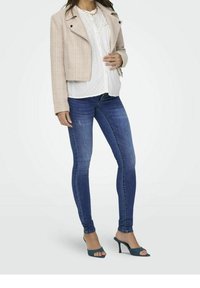 Chaqueta cropped texturizada rosa claro, blusa blanca con lunares, jeans pitillo azules y sandalias descubiertas teal. Collar de oro simple.