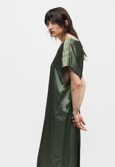 Άτομο με μακριά σκούρα μαλλιά και τατουάζ, φορώντας ένα φαρδύ, λαμπερό ελιάς πράσινο φόρεμα, στέκεται πλευρικά με τα χέρια δεμένα πίσω από την πλάτη.