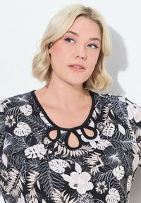 Svart blommig topp med korta ärmar, som har en detaljerad nyckelhålshalsringning. Materialet har en slät textur med vita blommor och bladmönster.