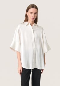 Camicia bianca in seta con vestibilità rilassata, dotata di maniche corte e ampie e colletto classico. Tessuto liscio, dettagli minimi.