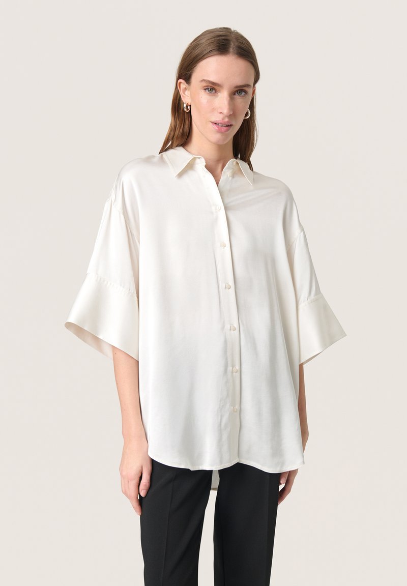 Camicia bianca in seta con vestibilità rilassata, dotata di maniche corte e ampie e colletto classico. Tessuto liscio, dettagli minimi.