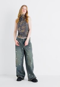 Top sem mangas com padrão camuflado, gola alta; jeans verdes largos com lantejoulas prateadas; tênis chunky pretos; fundo minimalista.