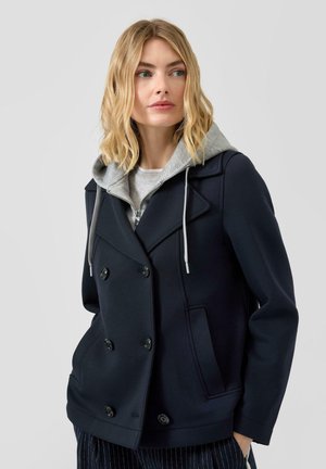 Blondynka w granatowym dwurzędowym płaszczu na szarej bluzie z kapturem i troczkami, patrząca nieco na bok na tle jednolitym.