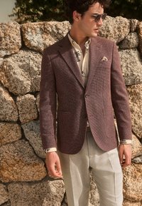Blazer en chevron marron avec une texture subtile, associé à une chemise légère à motifs floraux et à un pantalon gris clair ; posé devant un mur en pierre.