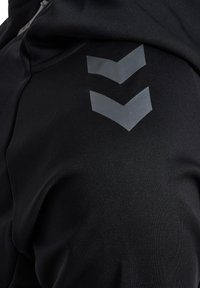Sort athletic stof med to glansfulde, grå chevronformer på siden. Teksturen er glat og strækbar, ideel til sportsbeklædning.