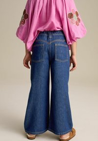 Vyöruhtaiset denim-housut tummassa pesussa, joissa on takataskut ja kontrastiväriset ompeleet. Yhdistetty vaaleanpunaisen brodeeratun pitkähihasen paidan kanssa.
