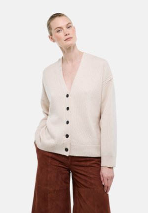 Femme portant un cardigan beige boutonné et un pantalon large marron, debout avec une main dans la poche devant un fond blanc uni.