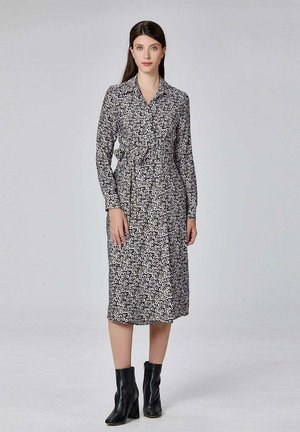 Une robe à manches longues et boutonnée présentant un motif floral noir et blanc, avec une taille froncée et tombant midi. Associée à des bottines noires.
