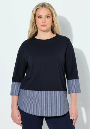 Kobieta z długimi blond włosami, ubrana w czarny top z paskowanymi, granatowo-białymi mankietami i dołem, stojąca na jednolitym jasnym tle.