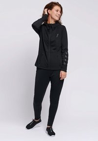 Czarna sportowa kurtka z zapięciem na zamek, kapturem i dwoma kieszeniami po bokach; czarne dopasowane spodnie; obie wykonane z gładkiego, elastycznego materiału.