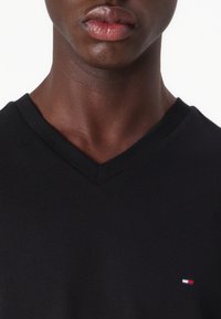 Krupan prikaz osobe koja nosi crnu majicu s V-izrezom i malim crveno-bijelo-plavim pravokutnim logotipom na prsima.