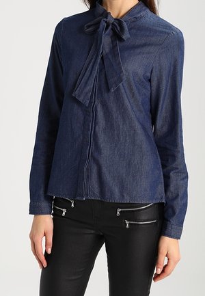 Femme portant un chemisier en jean à manches longues avec un nœud noué au col et un pantalon noir avec plusieurs poches zippées à l'avant.