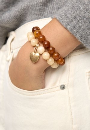 Nadgarstek z dwoma bransoletkami z koralików, jedna kremowa z złotym sercowym zawieszkiem, a druga w bursztynowym odcieniu, spoczywa w jasnokremowej kieszeni.