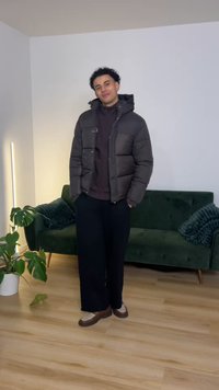 Geacă matlasată închisă la culoare cu guler înalt, pulover negru dedesubt, pantaloni largi negri și pantofi maro. Stă într-o cameră luminoasă cu o canapea verde.