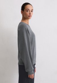 Szara bluza z okrągłym dekoltem, ściągaczami na mankietach i dole oraz miękką teksturą, noszona na ciemniejszym materiale pod spodem.