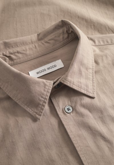 Camisa beige con cuello hecha de tela texturizada, que presenta una etiqueta con la inscripción "WOOD WOOD" y dos botones grises en la tapeta.
