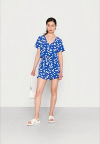 Macacão floral azul com mangas curtas, decote em V, cinto ajustável na cintura e design de calções de corte solto. Usado com sandálias brancas de slide.