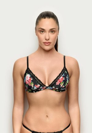 Mujer con cabello largo y oscuro, lleva un sujetador negro con encaje floral y ropa interior a juego, de pie contra un fondo claro y sencillo, mirando hacia adelante.