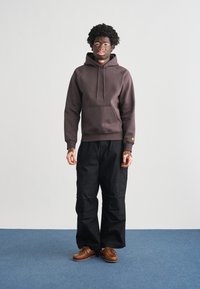 Καφέ φούτερ με κουκούλα, μπροστινή τσέπη και κορδόνι. Μαύρο παντελόνι τύπου cargo με πλαϊνές τσέπες. Καφέ παπούτσια. Απλός, καθημερινός σχεδιασμός.