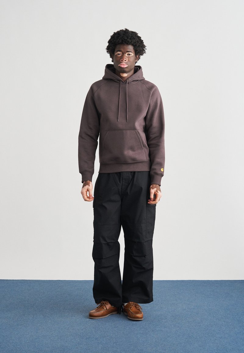 Brun hættetrøje med forlomme og snøre. Sorte cargobukser med sidelommer. Brune sko. Simpelt, afslappet design.