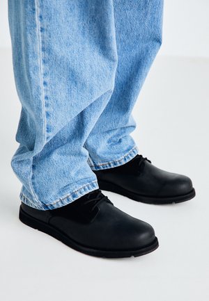 Chaussures noires en cuir avec un bout rond, à lacets, assorties à un jean en denim bleu clair avec des ourlets retroussés. Texture lisse, style simple.