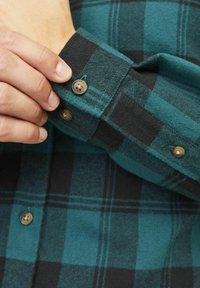 Koszula flanelowa w kratę w kolorze teal i czarnym, z zapięciem na guzik z złotymi guzikami, teksturowanym materiałem i kontrastującymi kolorami.