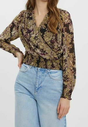 Kvinde iført en bluse med lange ærmer og blomstermønster med smocktalje og lyseblå, højtaljede denimjeans, stående mod en ensfarvet hvid baggrund.